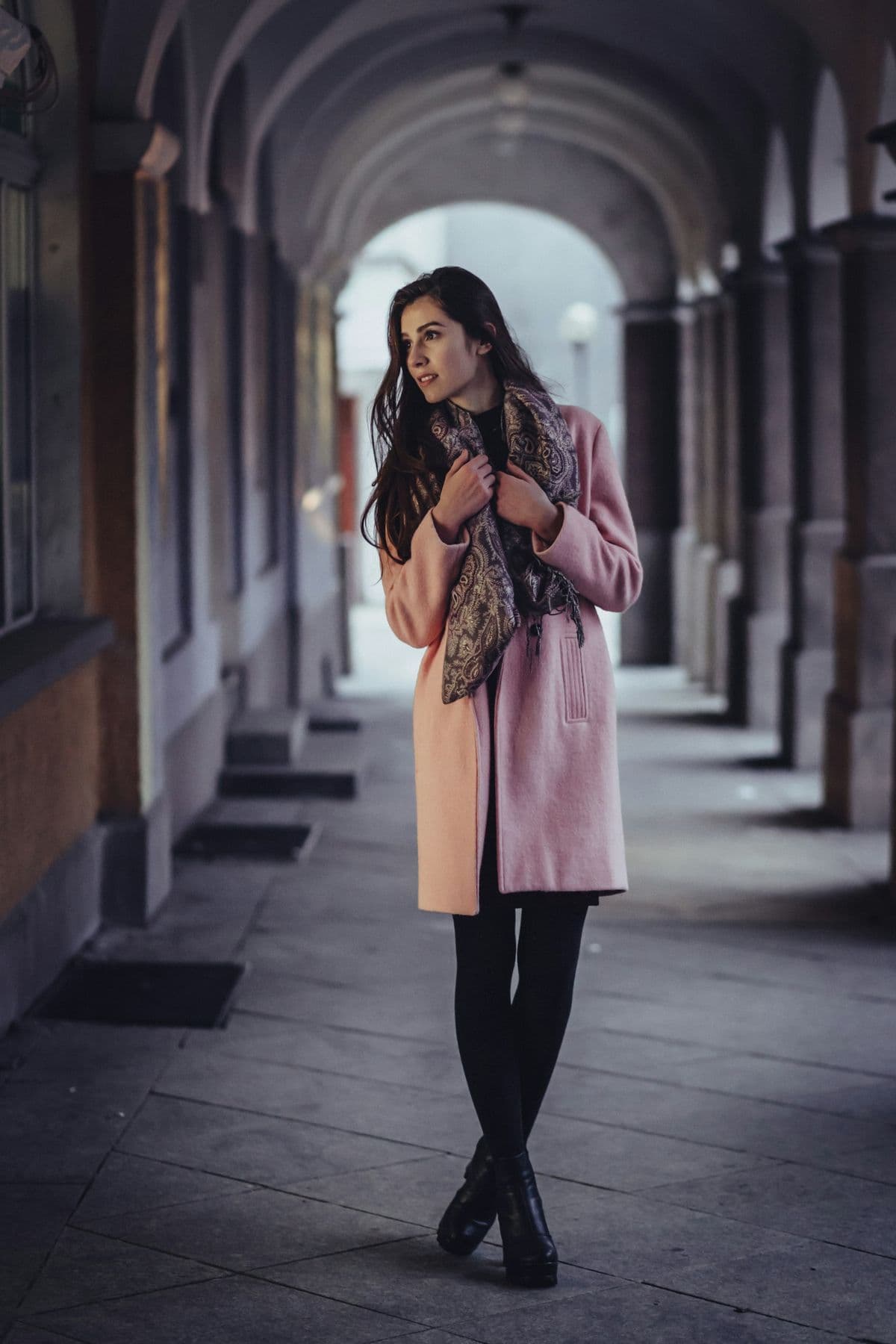 Woman in a dusty-pink wool coat standing beneath a stone arcade