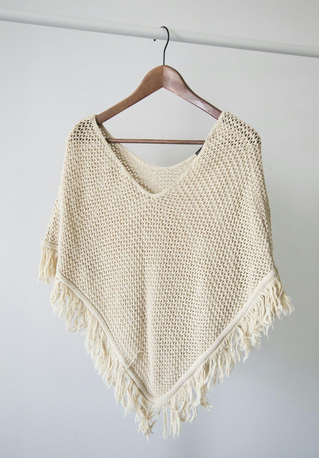 Cream knitted poncho on a wooden hanger against a white wall